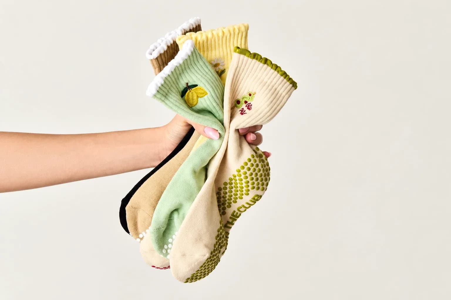 Een hand met een bosje gripsokken met ruches in mint, botergeel, beige en crème, met geborduurde details en een gestippelde gripzool